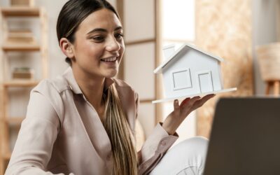 A woman working on her computer and holding a model of a house.